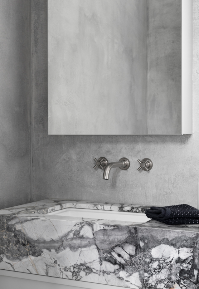 Contemporary pewter crosshead taps and tubular spout are wall mounted on mottled grey wall above solid marble basin vanity .